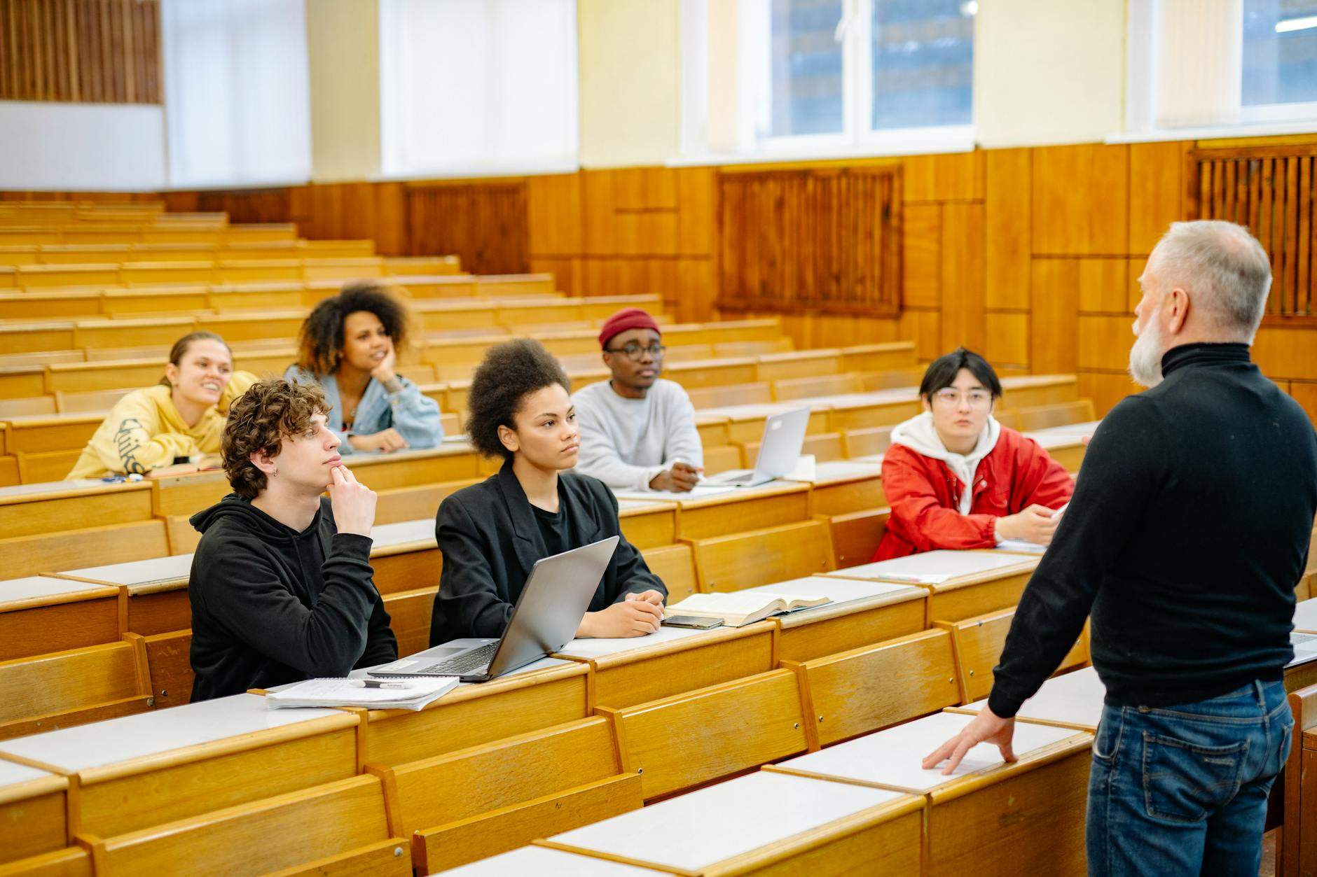 Grupa uczniów siedzących w sali wykładowej, słuchających prowadzącego stojącego przy pierwszym rzędzie ławek. Na stolikach widoczne są laptopy, zeszyty i podręczniki, a uczestnicy zajęć koncentrują się na omawianym materiale. Ujęcie nawiązuje do przygotow